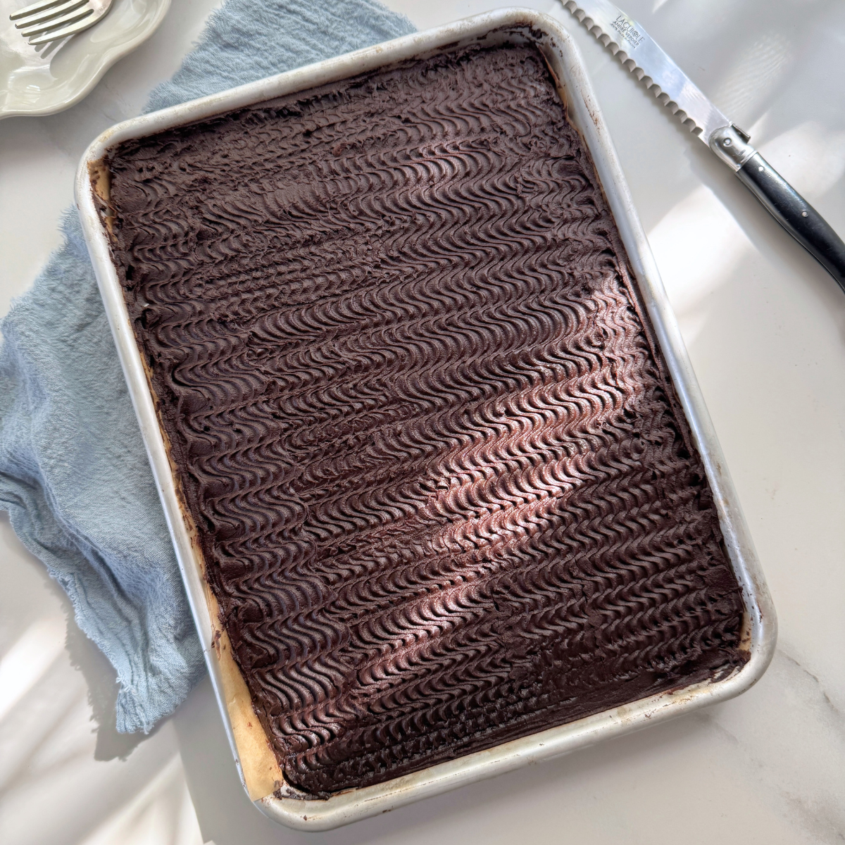 Full chocolate sheet cake in pan, topped with swirled buttermilk cocoa frosting using a comb for a wave pattern.