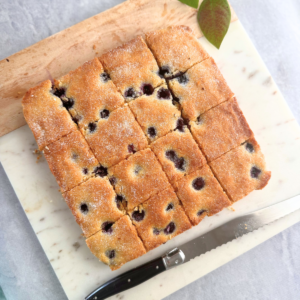Overhead view of soft, golden-baked blueberry yogurt bars cut into neat squares on a marble board, with a serrated knife beside them.