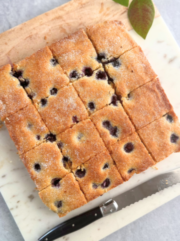 Overhead view of soft, golden-baked blueberry yogurt bars cut into neat squares on a marble board, with a serrated knife beside them.