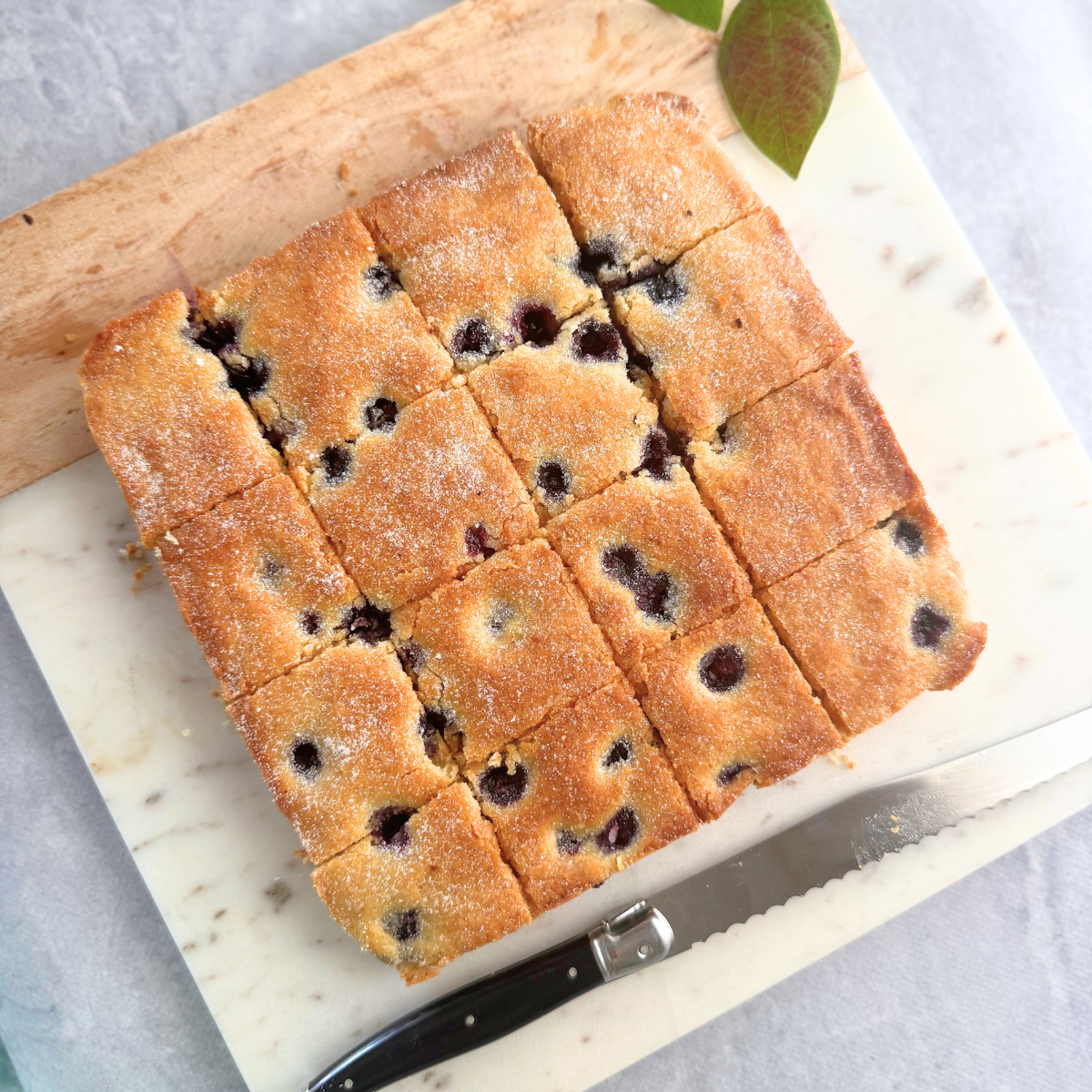 Overhead view of soft, golden-baked blueberry yogurt bars cut into neat squares on a marble board, with a serrated knife beside them.