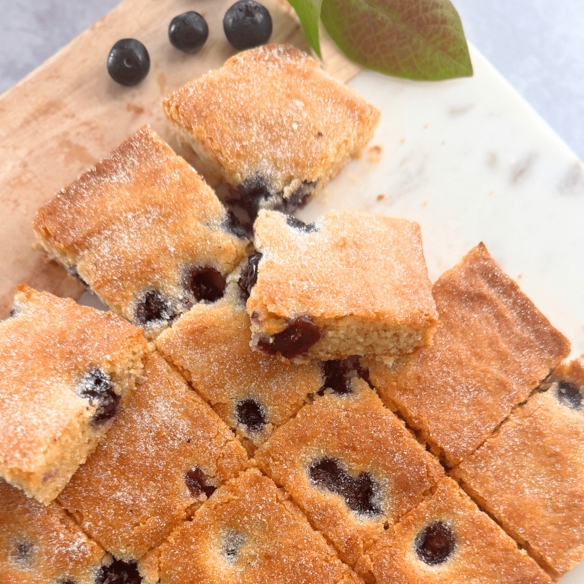 Golden-baked blueberry bars cut into squares, with a light sugar dusting on top. Some squares are slightly lifted to show the soft, moist interior filled with juicy blueberries. A few fresh blueberries and green leaves are placed nearby on a wooden and marble board.