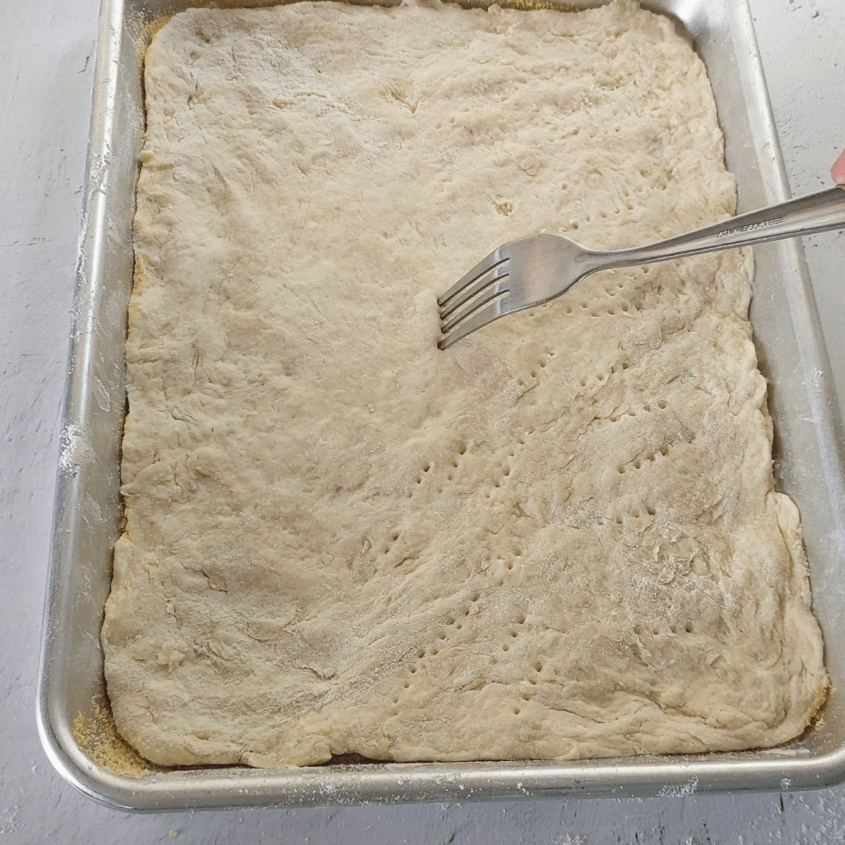 Unbaked yogurt-based pizza dough pressed into a rectangular sheet pan and being pricked with a fork to prevent bubbling.
