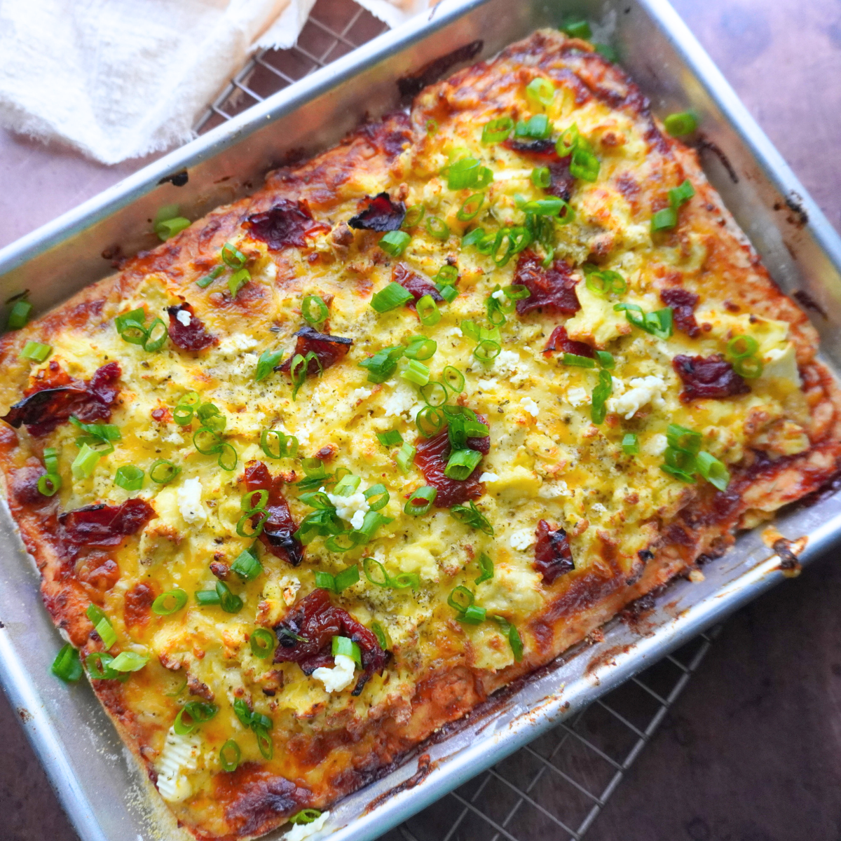 Fully baked breakfast pizza topped with scrambled eggs, sundried tomatoes, feta, and chopped green onions in a silver sheet pan on a cooling rack.