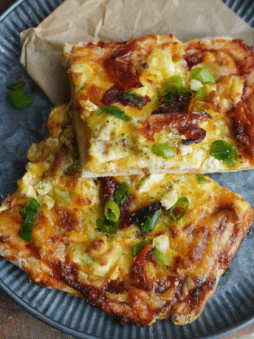 Two square slices of breakfast pizza with golden cheese, eggs, sundried tomatoes, and green onions, served on a gray plate with parchment paper.