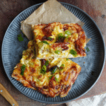 Two square slices of breakfast pizza with golden cheese, eggs, sundried tomatoes, and green onions, served on a gray plate with parchment paper.