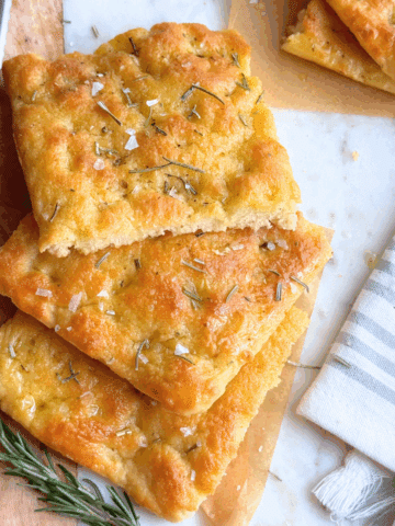 Top view of sliced no-yeast focaccia topped with rosemary and flaky salt, arranged on parchment with a serrated knife and striped kitchen towel