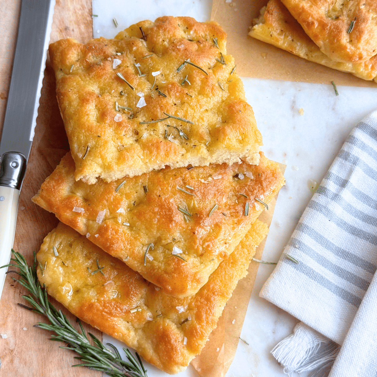 Top view of sliced no-yeast focaccia topped with rosemary and flaky salt, arranged on parchment with a serrated knife and striped kitchen towel