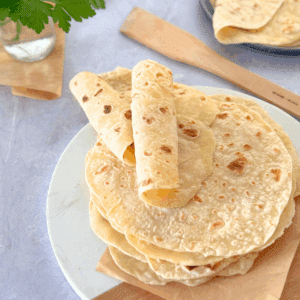 Soft, golden brown cottage cheese flatbreads stacked on a wooden board, with one rolled and ready to serve.
