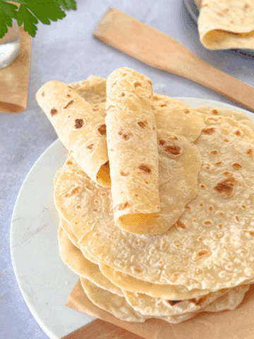 Soft, golden brown cottage cheese flatbreads stacked on a wooden board, with one rolled and ready to serve.
