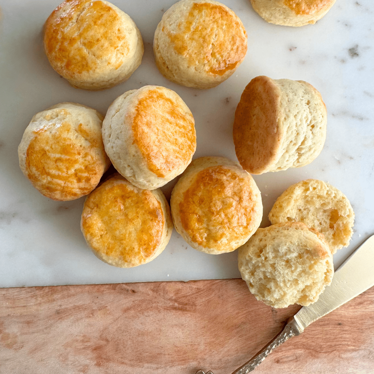 Mini cream scones with golden tops, one split open to show a soft, fluffy inside, on a marble board with a butter knife.