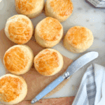 Golden mini cream scones arranged on a marble surface, served with jam and cream in the background.