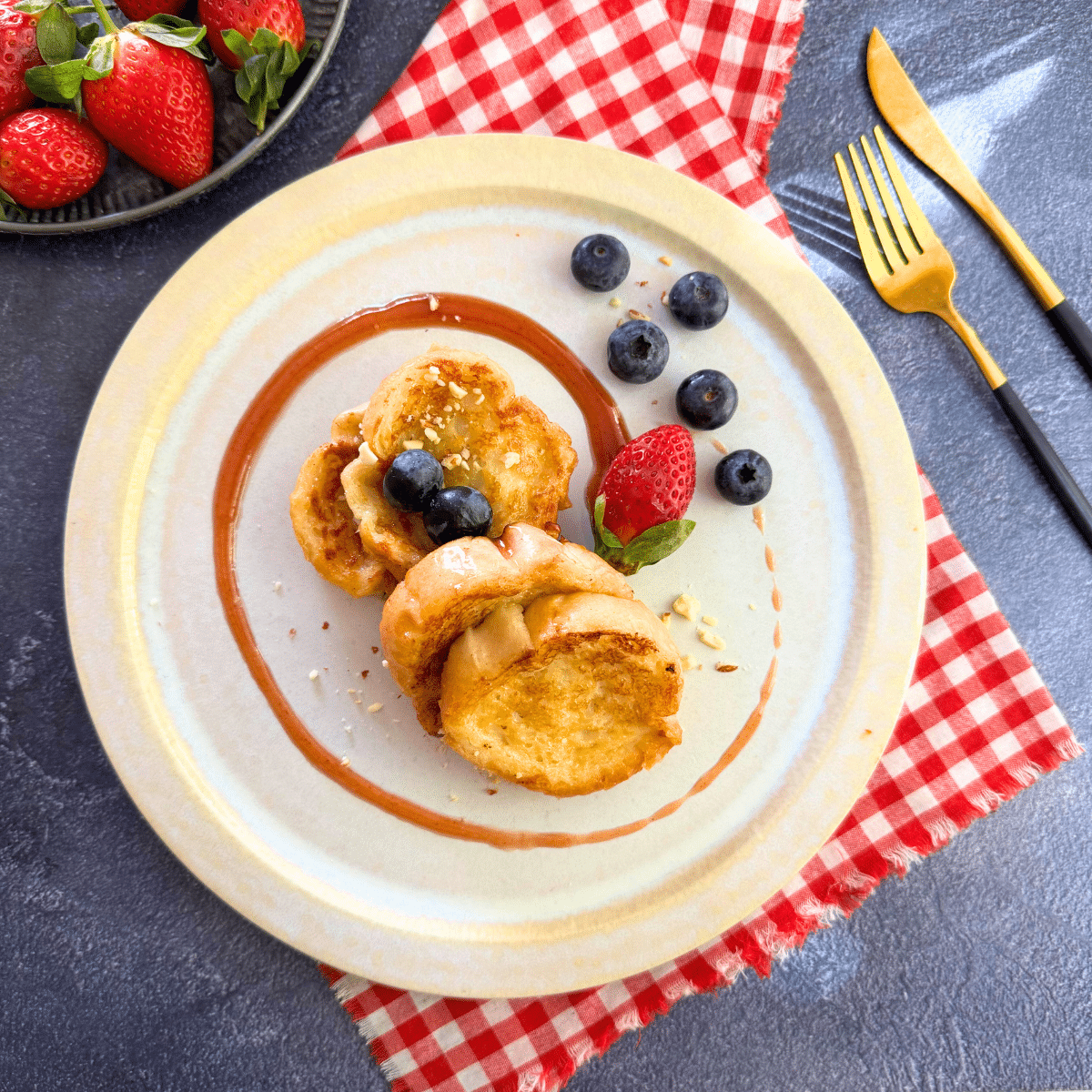 Overhead view of mini French toast made without eggs, plated with berries and a swirl of jam on a white dish with a red checkered napkin.