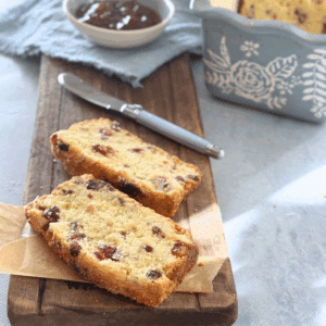 Angled view of cranberry scone loaf slices in front of the full loaf in a baking pan, with jam and knife beside