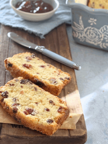 Angled view of cranberry scone loaf slices in front of the full loaf in a baking pan, with jam and knife beside