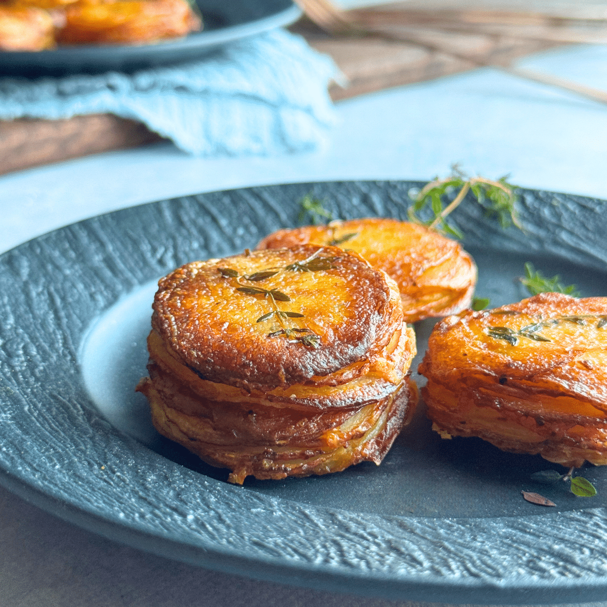 Crispy golden garlic butter potato stacks garnished with thyme, stacked neatly on a dark ceramic plate with a soft, rustic background