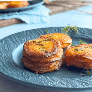 Crispy golden garlic butter potato stacks garnished with thyme, stacked neatly on a dark ceramic plate with a soft, rustic background