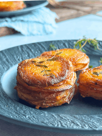 Crispy golden garlic butter potato stacks garnished with thyme, stacked neatly on a dark ceramic plate with a soft, rustic background