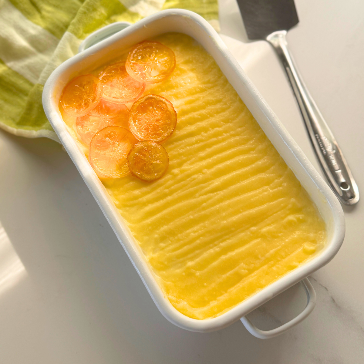 Overhead view of yogurt lemon tiramisu in a white rectangular dish, topped with lemon curd and glossy candied lemon slices.